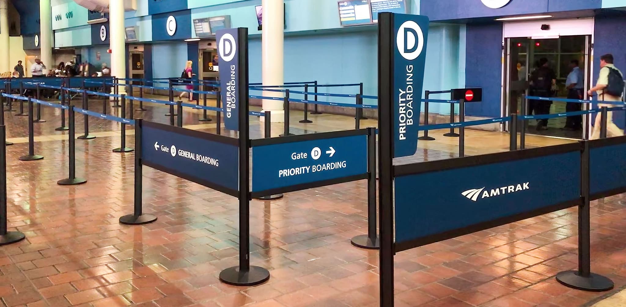 Partition posts with Rigid Rail barriers and banner signs form the boarding queue at an Amtrak station.